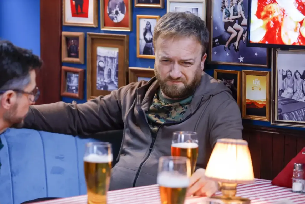 Tatusiowie serial obsada: Mężczyzna z brodą i w bluzie siedzi przy stole z piwem, w tle ściana z ramkami.