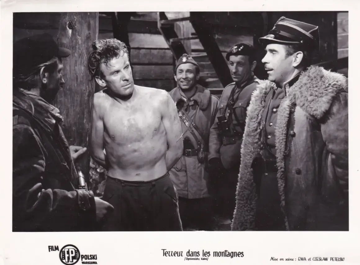 Czarno-białe zdjęcie przedstawia scenę z filmu: mężczyzna bez koszuli, z rękami związanymi, stoi przy drewnianym słupie, otoczony przez żołnierzy.