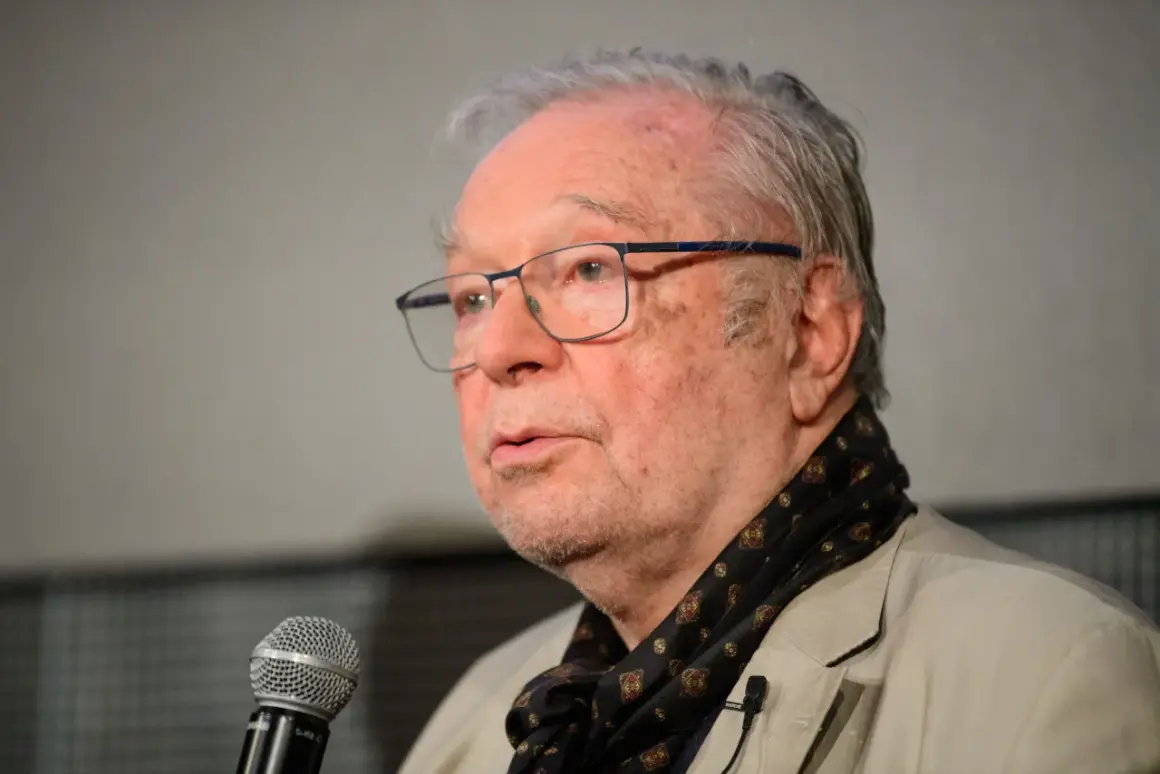 Portret starszego mężczyzny w okularach, mówiącego do mikrofonu, ubranego w jasną marynarkę i wzorzysty szalik.