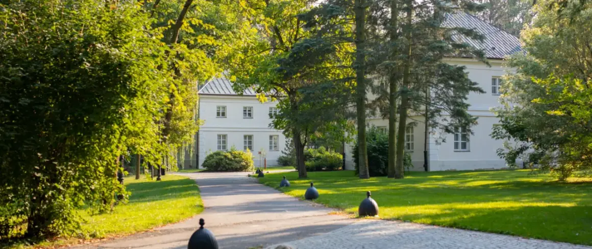 Biały dworek otoczony zielenią, z alejką i ozdobnymi kulami.