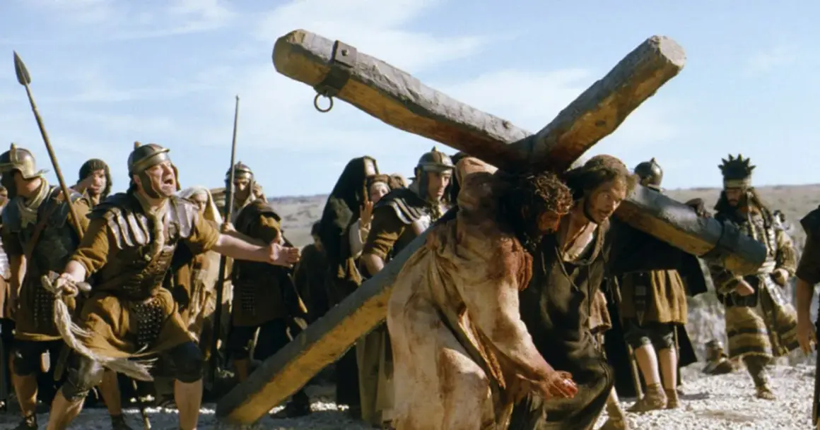 Jezus w koronie cierniowej niesie krzyż, otoczony przez rzymskich żołnierzy.