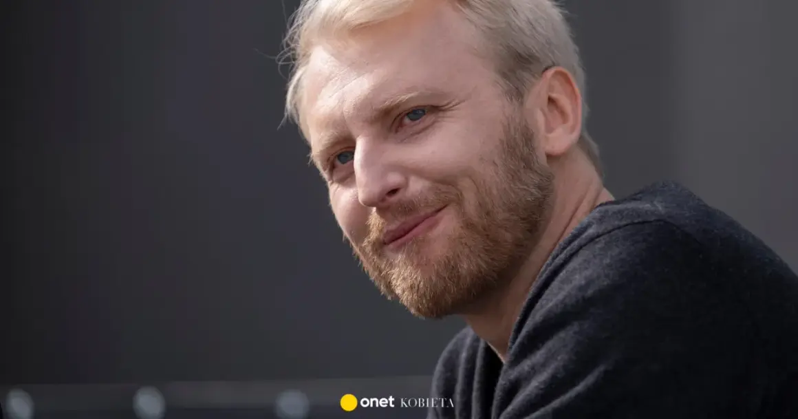 Uśmiechnięty Dobromir Dymecki z jasnymi włosami i niebieskimi oczami, w ciemnej bluzie.