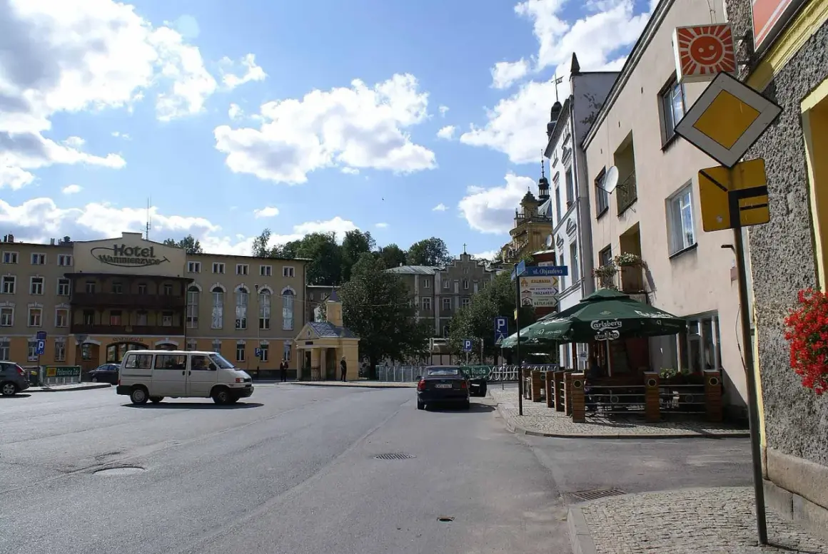 Plac miejski z hotelem, samochodami i kawiarnią pod zielonymi parasolami.