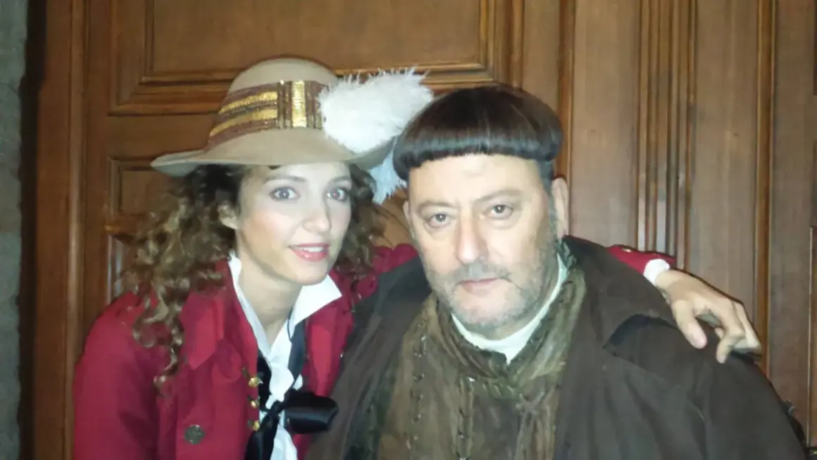 Goście, goście, obsada w historycznych strojach. Kobieta w kapeluszu z piórem i mężczyzna w brązowym płaszczu pozują do zdjęcia.