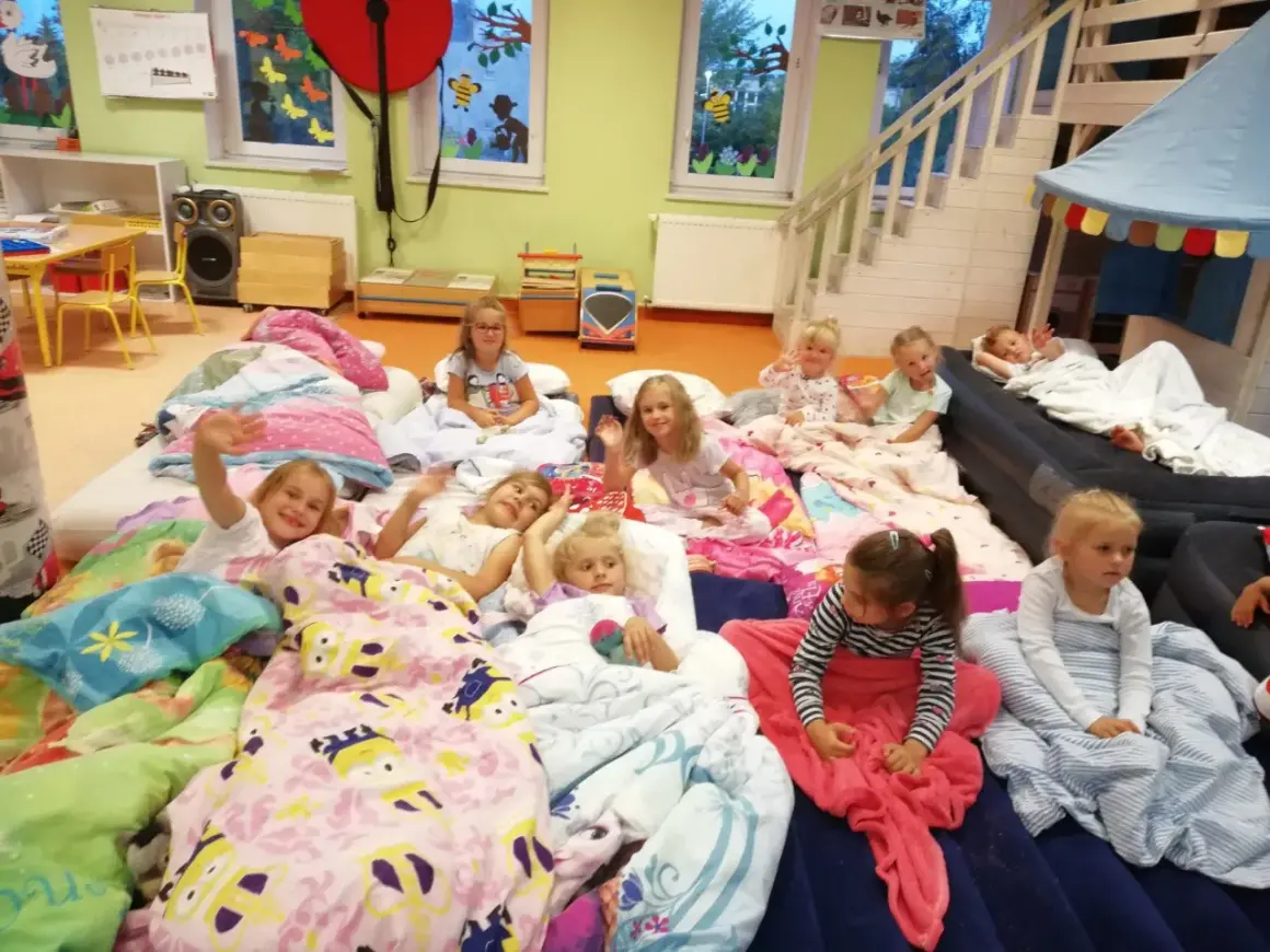 Dzieci leżą w śpiworach i kołdrach, gotowe na noc w przedszkolu. Niektóre machają do aparatu, inne patrzą z zaciekawieniem.