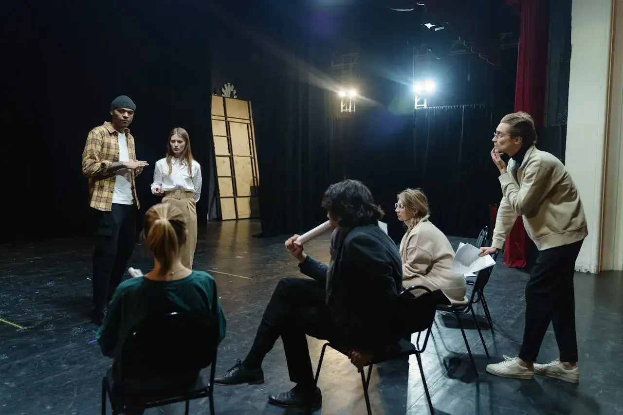 Grupa ludzi na scenie, dyskutująca o spektaklu. Zastanawiają się, ile zarabia reżyser, analizując scenariusz.