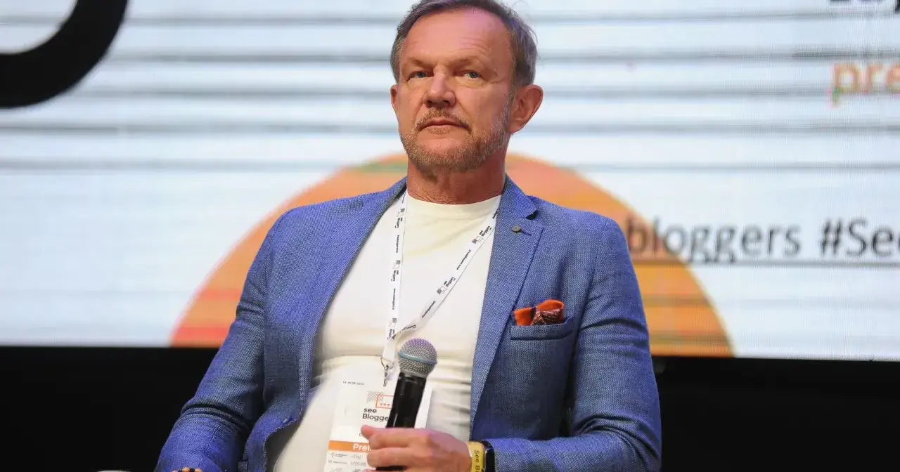 Mężczyzna w niebieskiej marynarce, trzymający mikrofon, podczas panelu dyskusyjnego. Ciekawe, ile zarabia Cezary Pazura, gdyby występował w podobnych wydarzeniach.