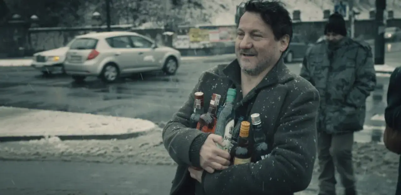 Mężczyzna w płaszczu, pod mocnym aniołem obsada, niesie kilka butelek alkoholu. Pada śnieg.