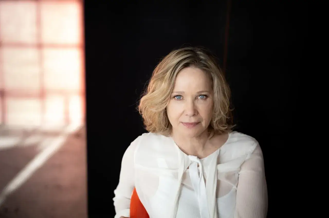 Portret Ann Kathrin Kramer z niebieskimi oczami, w białej bluzce, na tle czerni i rozmytego okna.