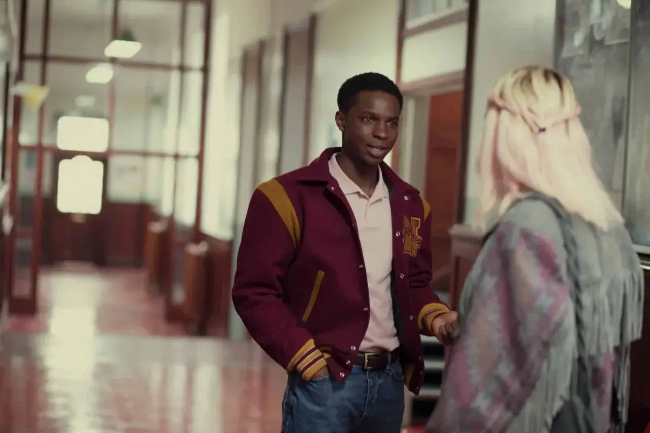 Kedar Williams Stirling w bordowej kurtce z żółtymi wstawkami rozmawia z koleżanką na szkolnym korytarzu.
