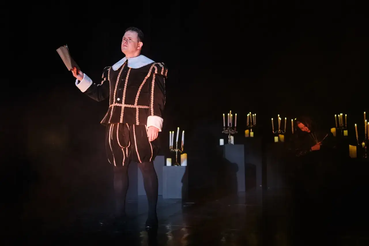 Aktor w stroju z epoki trzyma kartkę, zastanawiając się, ile zarabia aktor za spektakl. W tle płoną świece.