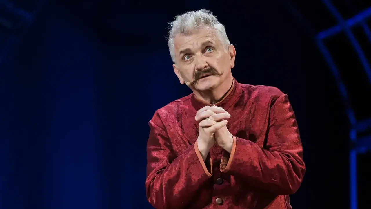 Dariusz Kowalski, aktor, w czerwonym stroju z charakterystycznym wąsem, z dłońmi złożonymi w geście błagania.
