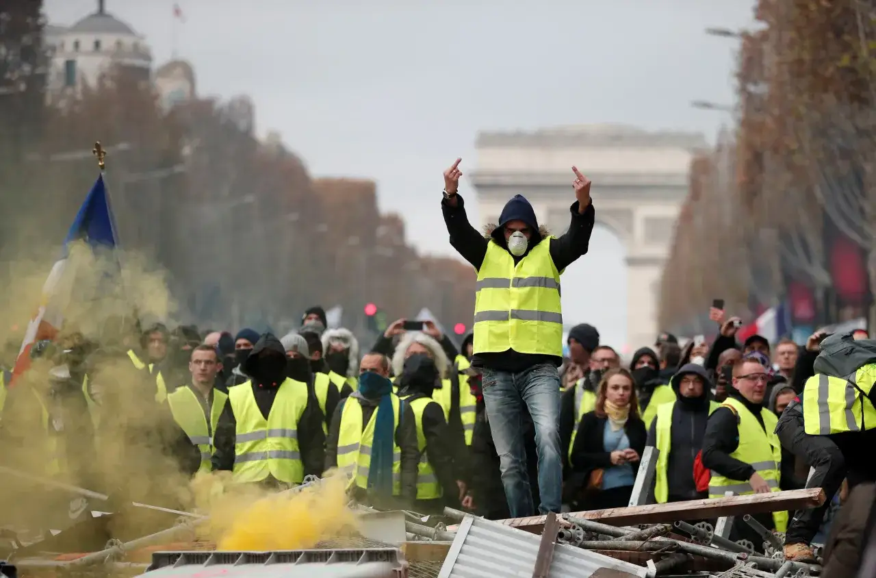 Paryż w ogniu! Protestujący w kamizelkach odblaskowych, z flagą Francji i żółtym dymem, na tle Łuku Triumfalnego.