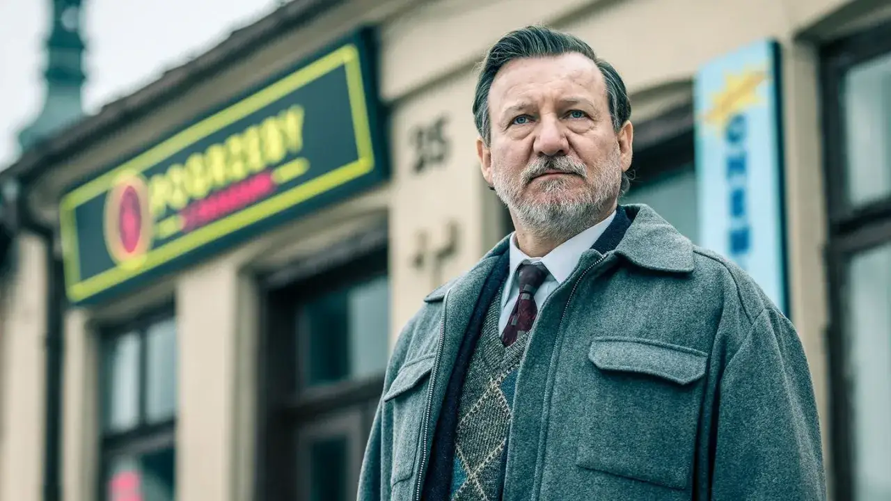 Mężczyzna w szarym płaszczu i swetrze w romby, stojący przed budynkiem z neonem. To część obsady serialu 