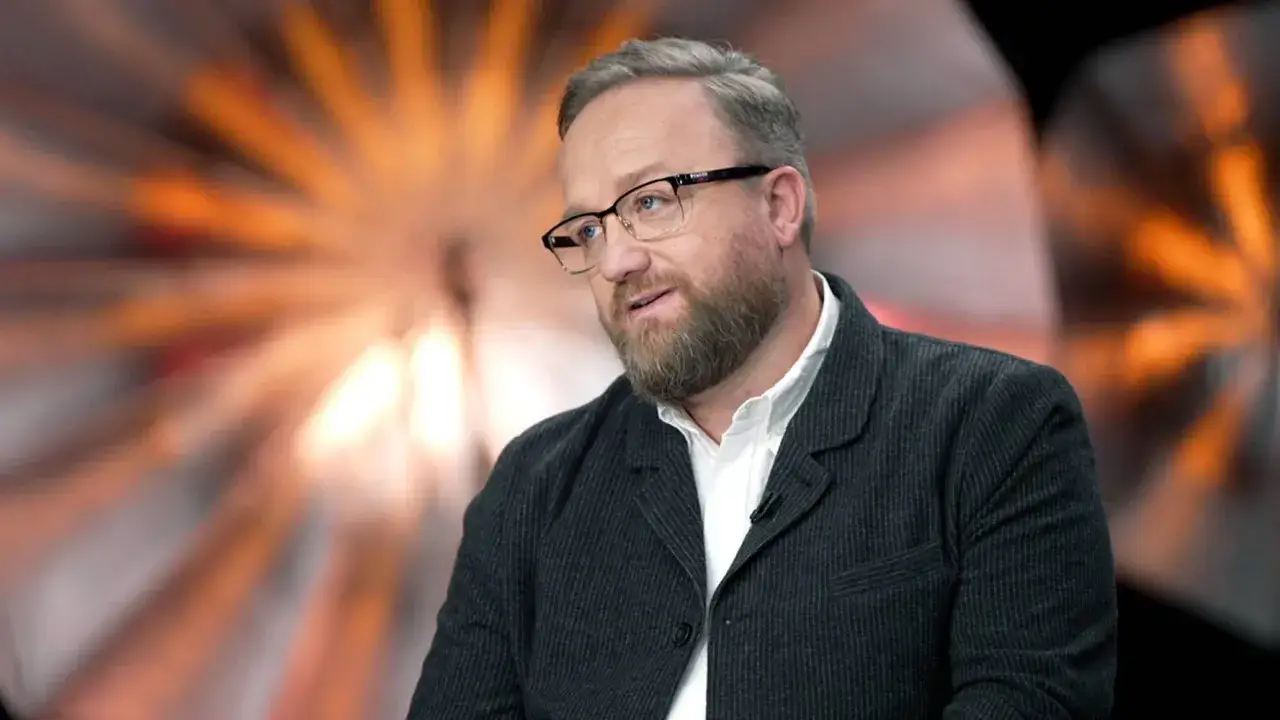 Bartłomiej Kasprzykowski w okularach i marynarce, z zarostem, na tle rozmytego, pomarańczowego światła.