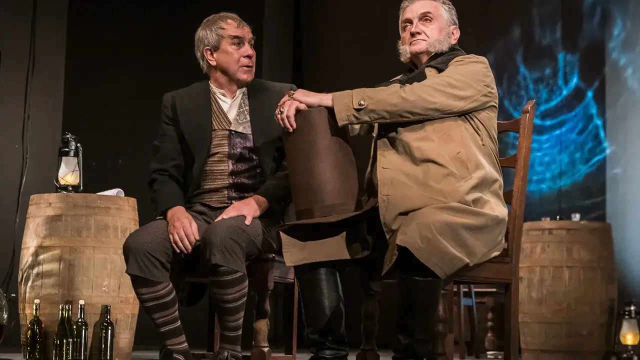 Dariusz Kowalski aktor w teatralnej scenografii, siedzi obok kolegi, trzymając kapelusz.
