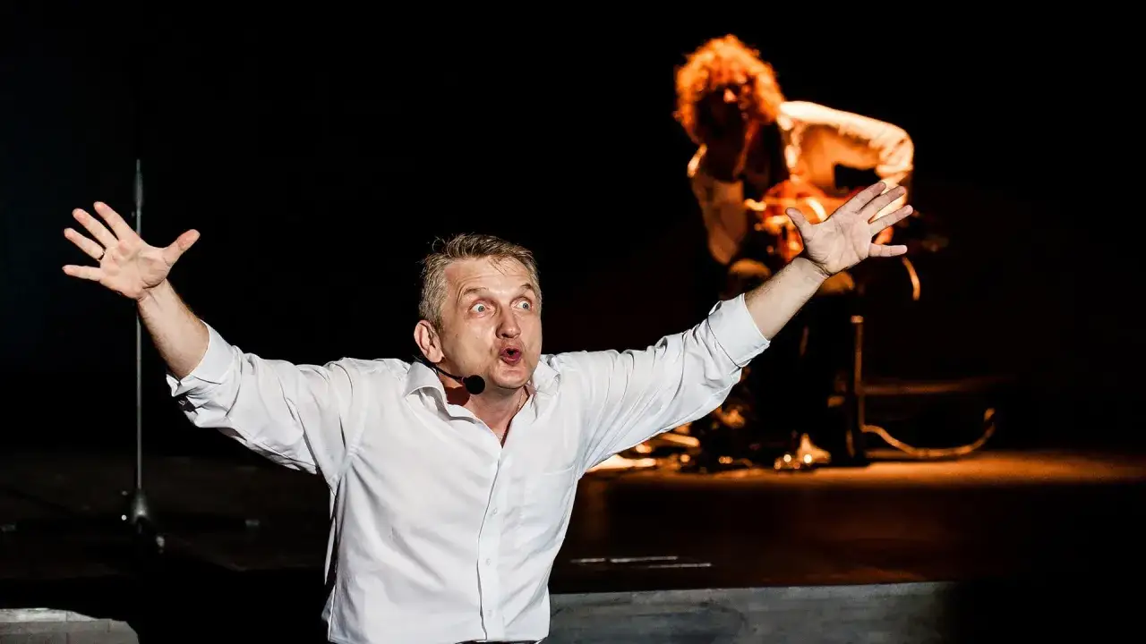 Dariusz Kowalski, aktor, z szeroko otwartymi ramionami i wykrzywionymi ustami, w białej koszuli, na scenie.