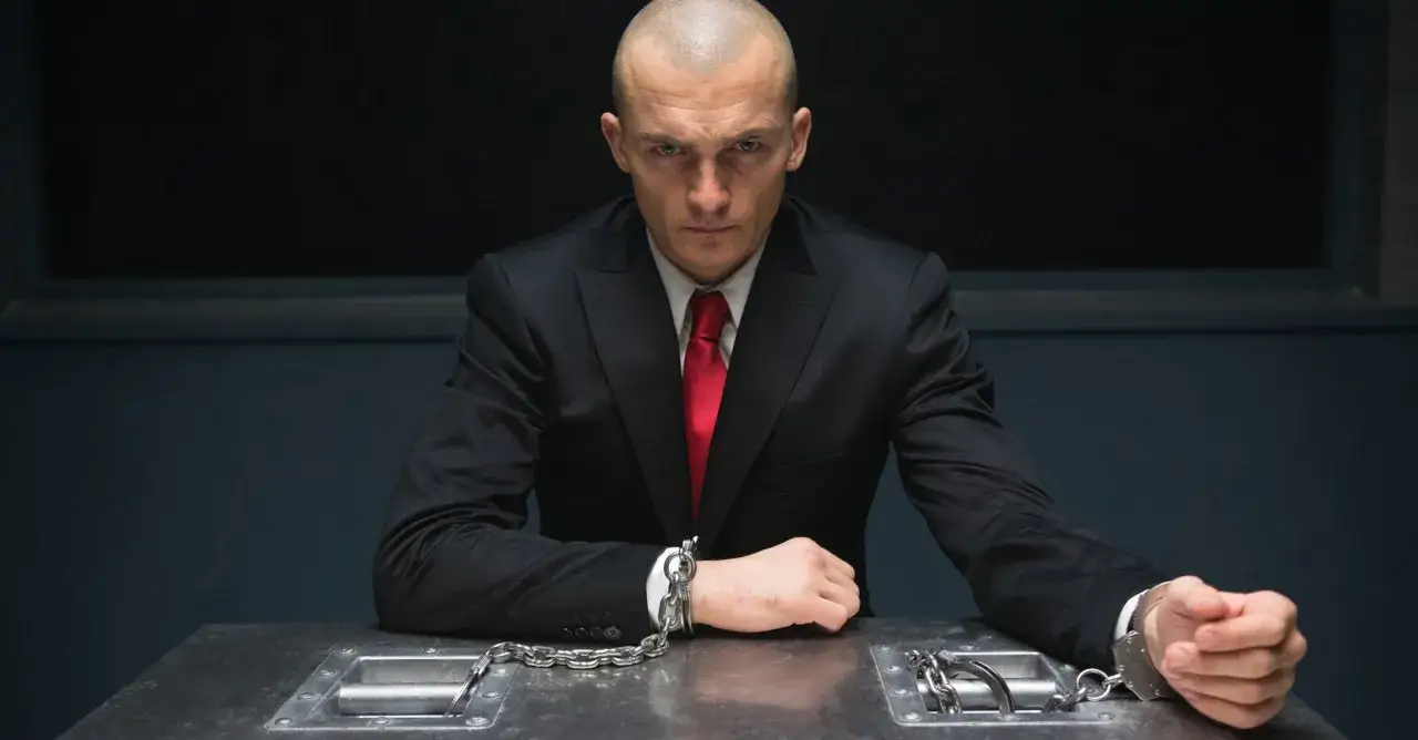 Agent 47 w garniturze i czerwonym krawacie, skuty kajdankami, siedzi przy stole.