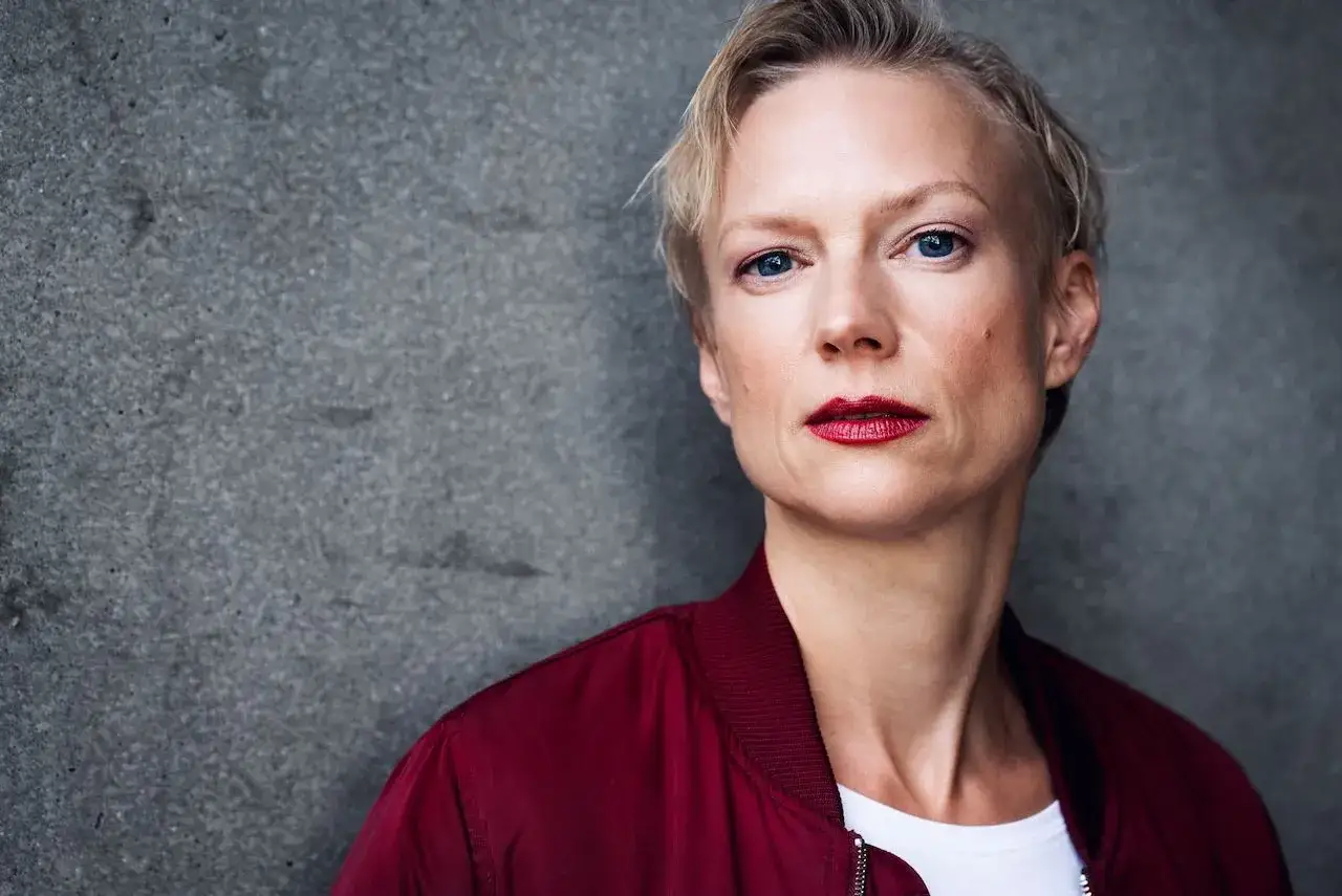 Portret Lise Risom Olsen w bordowej kurtce bomberce, z wyrazistymi czerwonymi ustami i niebieskimi oczami, na tle szarej ściany.