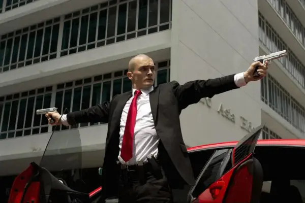 Agent 47 w czarnym garniturze i czerwonym krawacie, z dwoma pistoletami w dłoniach, gotowy do akcji.