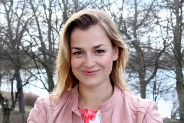 Uśmiechnięta Ines Lutz, znana z roli w "Der Bergdoktor", w różowej kurtce na tle drzew.
