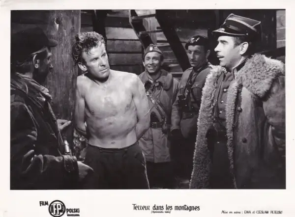 Czarno-białe zdjęcie przedstawia scenę z filmu: mężczyzna bez koszuli, z rękami związanymi, stoi przy drewnianym słupie, otoczony przez żołnierzy.