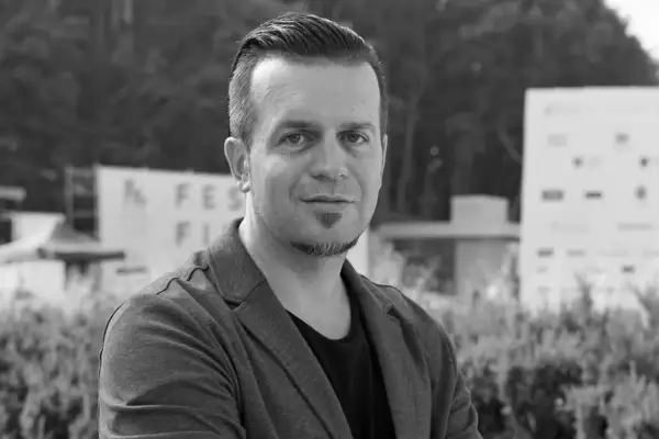 Czarno-białe zdjęcie Marcina Wrony, reżysera, w marynarce, na tle budynków i zieleni.