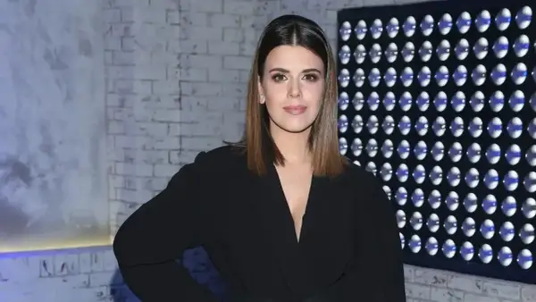 Monika Mazur w czarnym eleganckim stroju pozuje na tle ceglanej ściany i panelu z niebieskimi światłami.