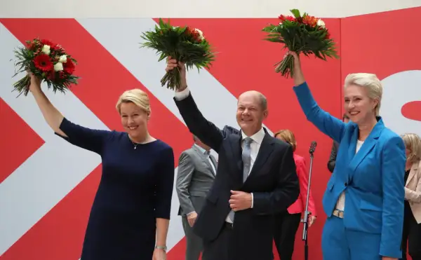 Uśmiechnięta Astrid Ann Marie Pollmann i politycy z bukietami kwiatów na tle czerwono-białej ściany.