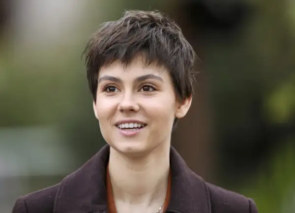 Uśmiechnięta aktorka na dobre i na złe, z krótkimi, ciemnymi włosami i w brązowym płaszczu.