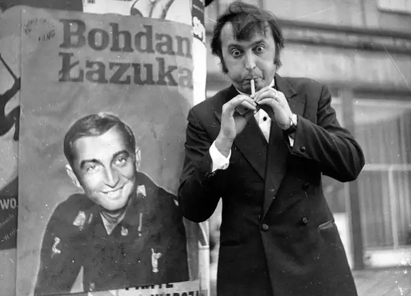 Bohdan Łazuka w garniturze, z papierosem w ustach, obok plakatu z jego podobizną.