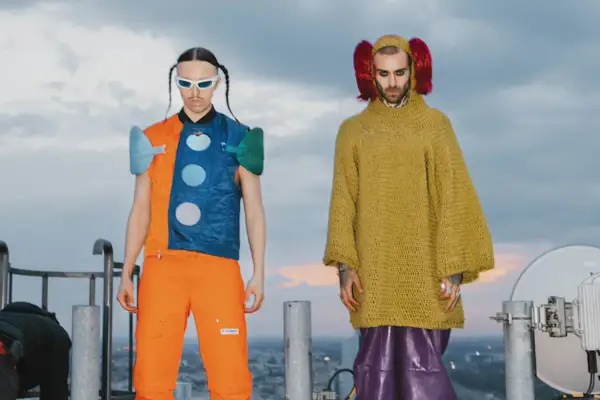 Tommy Cash i Quebonafide w ekstrawaganckich strojach na dachu. Jeden w pomarańczowo-niebieskiej kamizelce, drugi w żółtym swetrze i fioletowych spodniach.