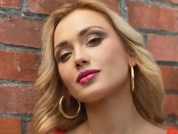 Dominika Skoczylas z błyszczącymi złotymi kolczykami i wyrazistym makijażem ust, na tle ceglanej ściany.