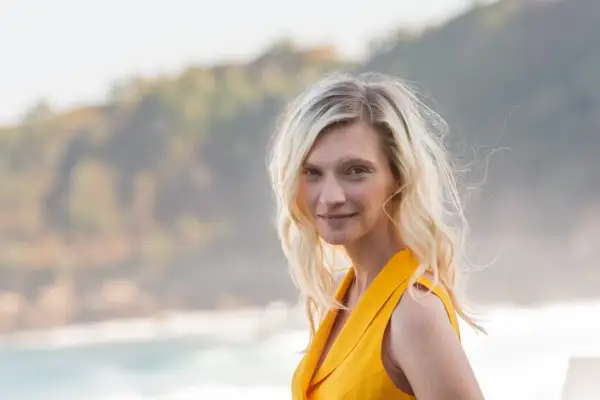 Agata Buzek w żółtej sukience, z falującymi blond włosami, uśmiecha się na tle morza i wzgórz.